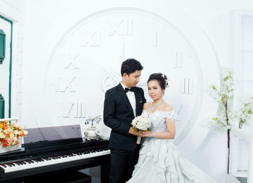 Asian couple in elegant wedding attire posing near a grand piano with floral decor.