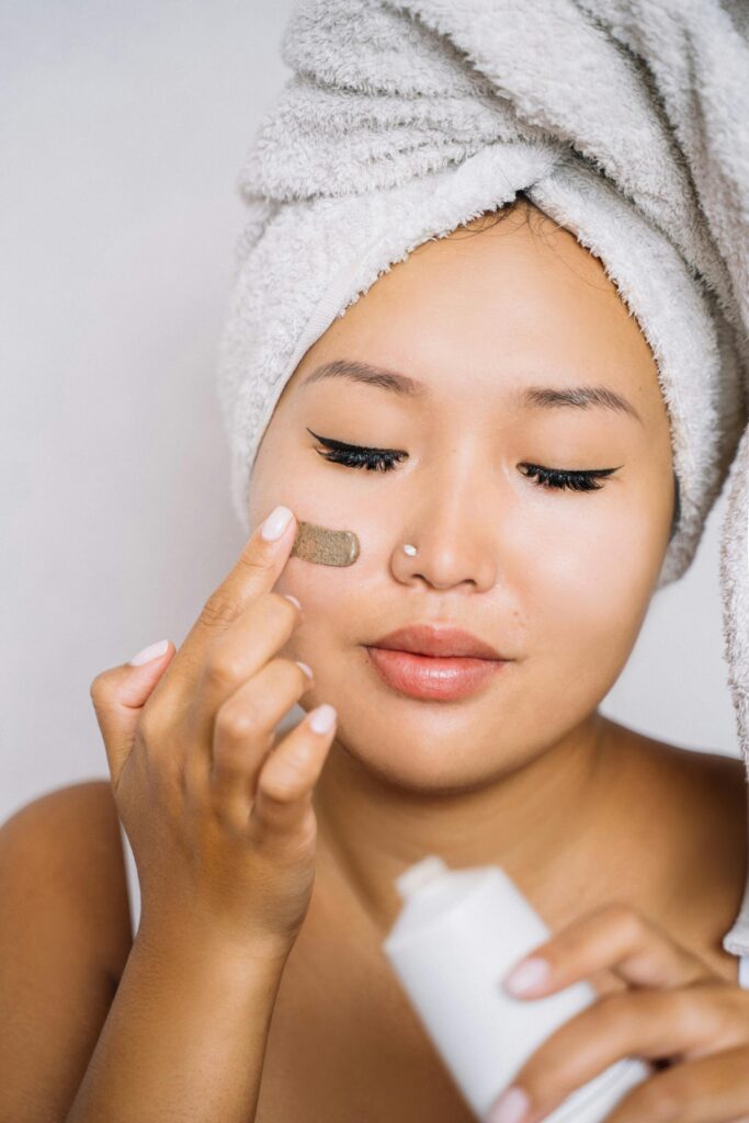 Asian woman with towel headwrap applying facial cream, focusing on skincare routine.