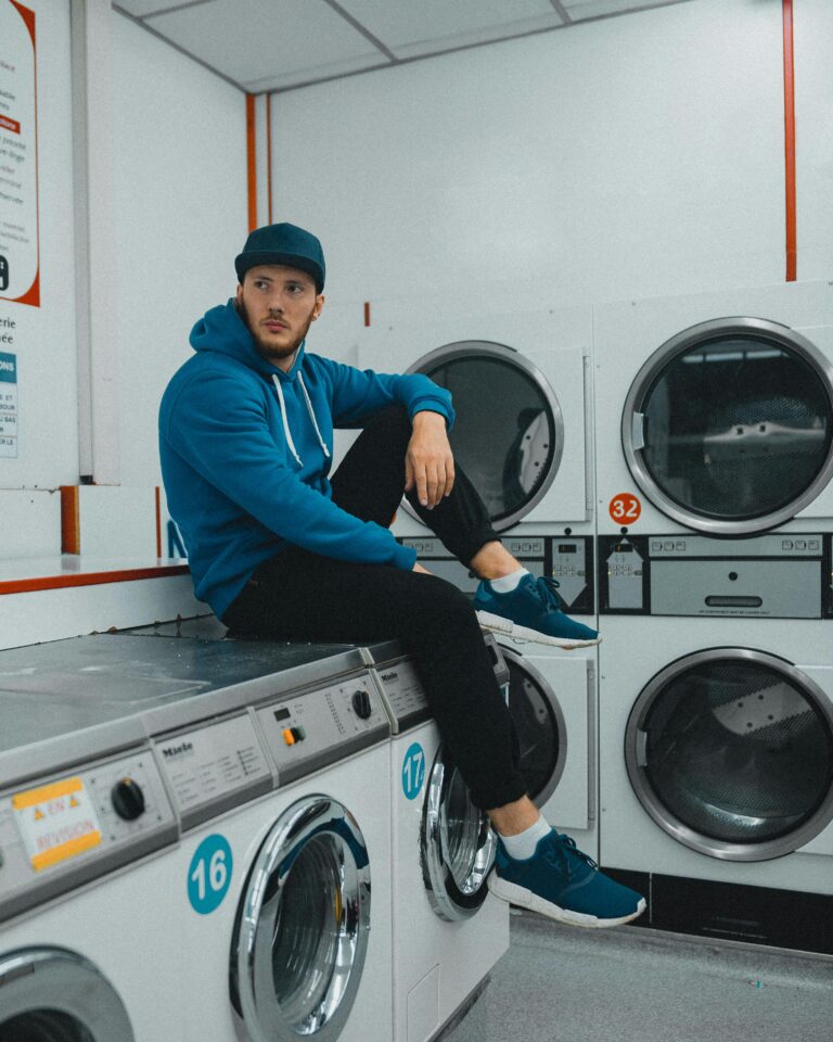 A man casually sitting in a laundromat, wearing a hoodie and cap, with washing machines in view.